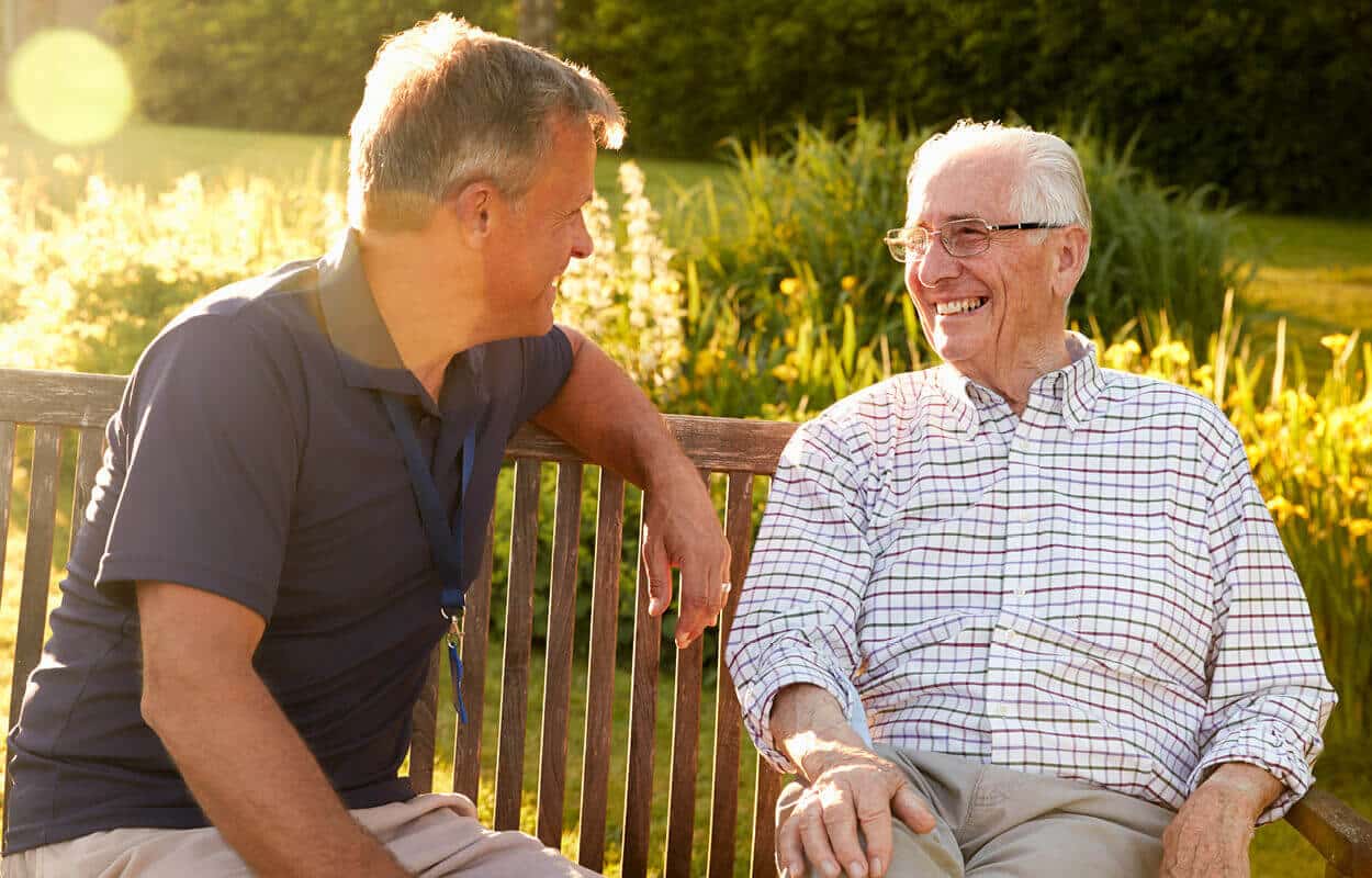 A senior man and middle-aged man smiling and sitting on park bench