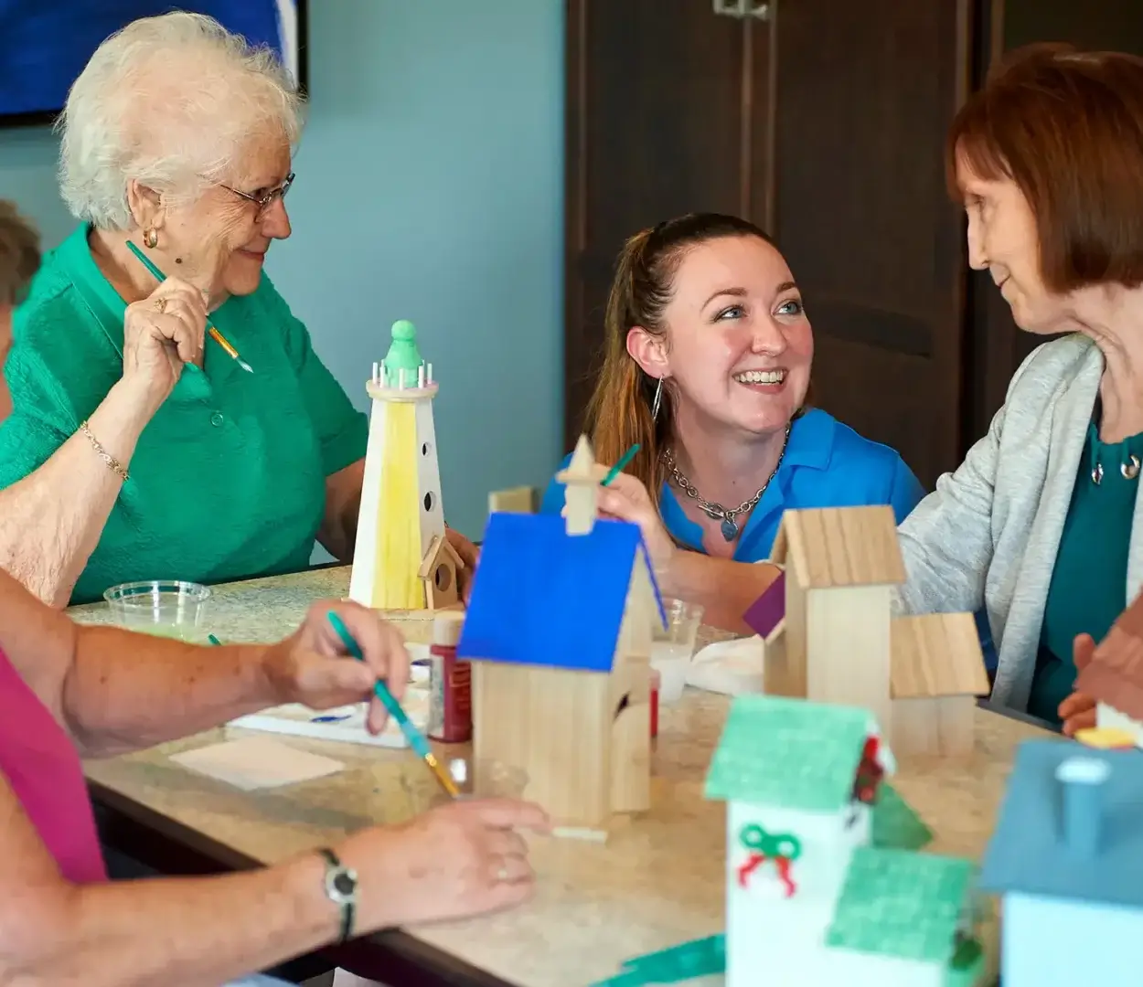 three senior resident women painting birdhouses in art room, with associate woman kneeling next to table